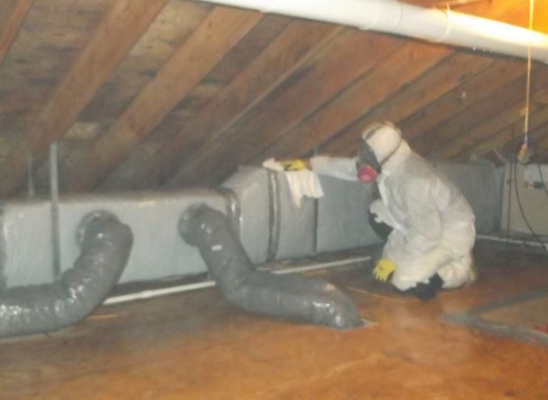 Worker in a protective suit and respirator treating attic framing near HVAC ducts